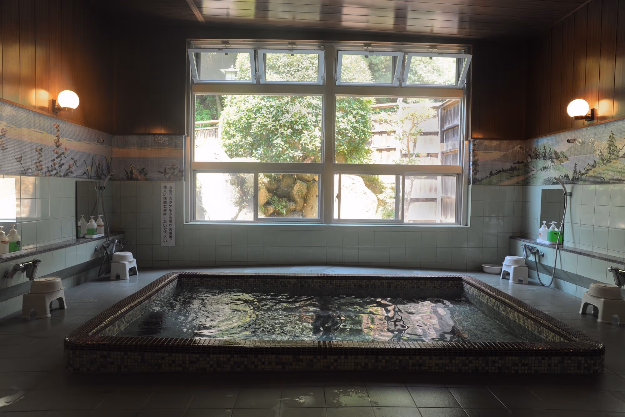 Indoor Japanese onsen bath with tiled walls, stools, shower heads, and a large window showing greenery outside.