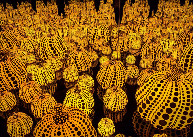 Multiple yellow pumpkins covered in black polka dots reflected infinitely in mirrors, creating a pattern of repetitive pumpkin shapes.