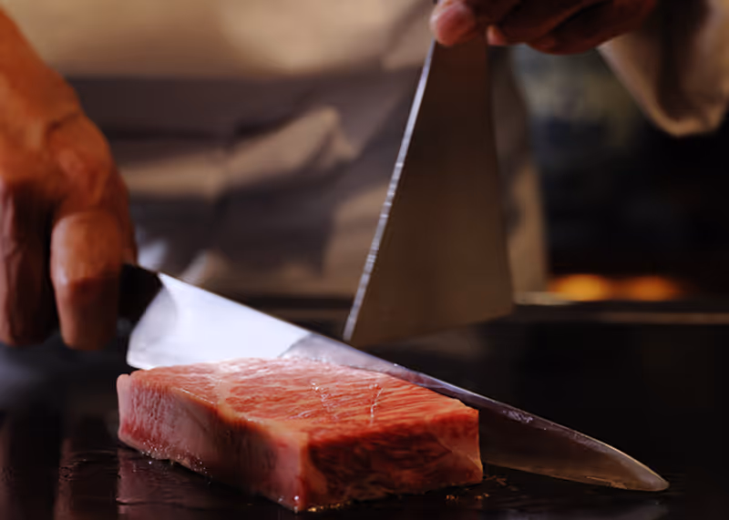 Chef slicing a thick piece of raw marbled beef with a large knife on a dark surface.