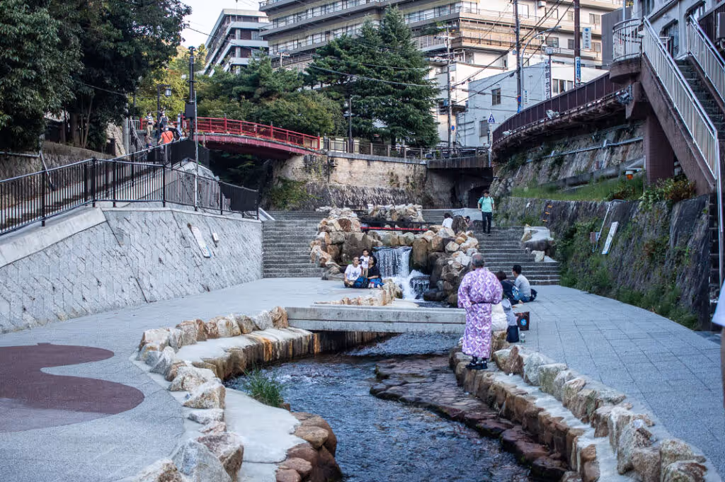 People enjoy a stone-lined river with small waterfalls in an urban park, including a person wearing a purple kimono, near a red bridge and multi-story buildings.