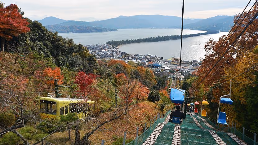 A scenic view of a chairlift and cable car over autumn foliage overlooking a coastal town and a long sandbar extending into the water with mountains in the distance.
