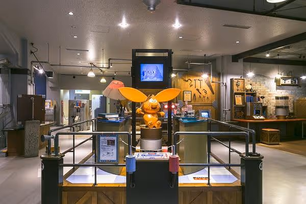 Interior view of a children’s cultural and science museum exhibit featuring a bright orange animated character with large ears on a pedestal, surrounded by interactive displays and screens.