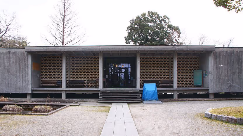 Front view of a minimalist concrete building with lattice brick walls and a central entrance, surrounded by sparse trees and a gravel pathway.