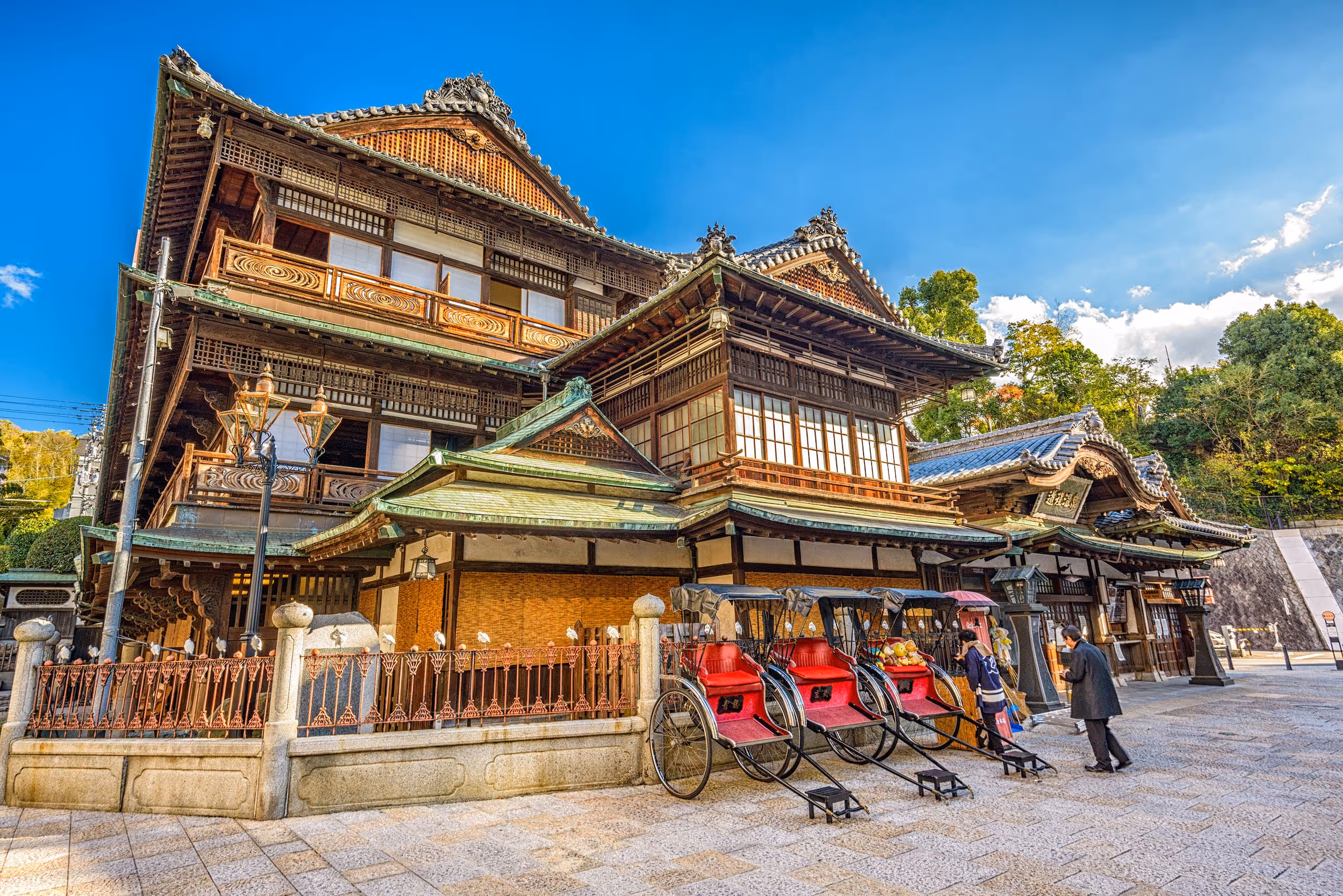 Historic multi-story traditional Japanese wooden building with ornate roof and three red rickshaws parked in front under a clear blue sky.