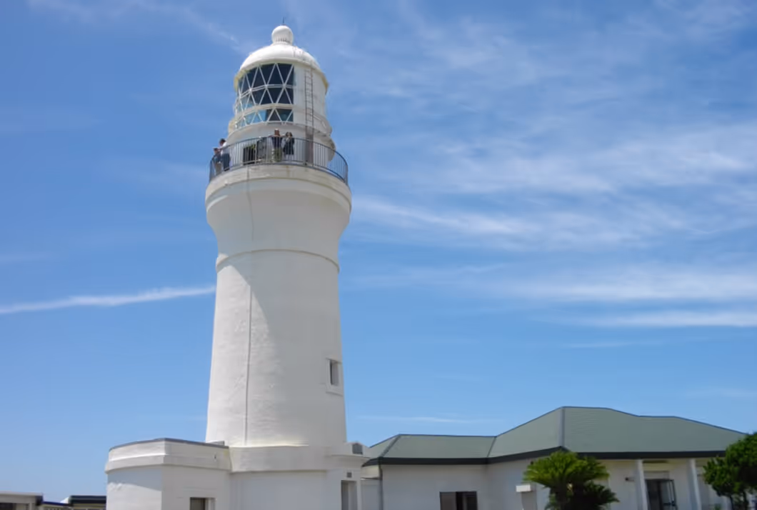 White lighthouse with people on the balcony against a blue sky with scattered clouds.
