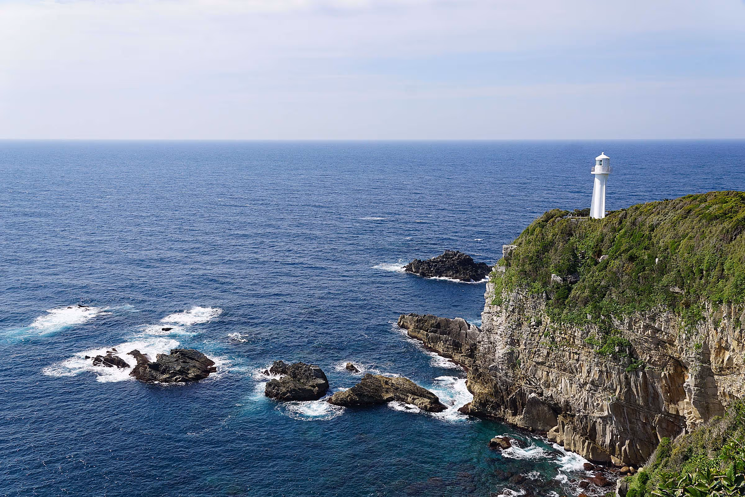 White lighthouse on a green cliff overlooking a rocky coastline and blue ocean under a clear sky.