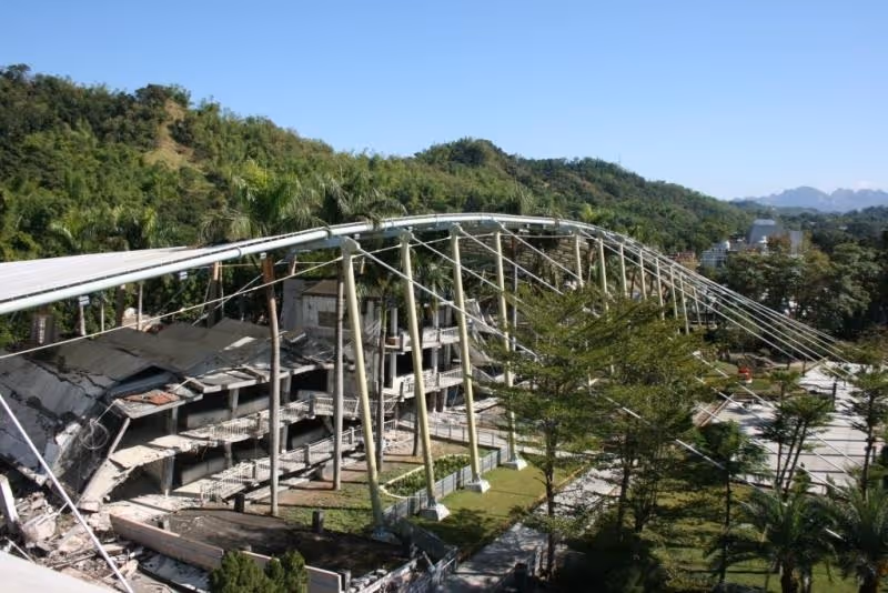 Partially collapsed multi-story concrete building supported by large metal poles and cables in a green mountainous area under blue sky.