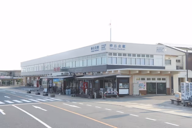 Modern two-story building with large windows labeled 'Sakyu Kaikan' and 'Sakyu Kaikan' in Japanese, situated next to a wide road with marked lanes.