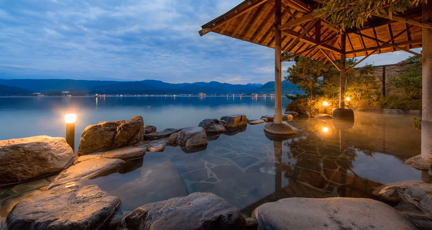 Outdoor stone hot spring bath with wooden shelter overlooking a calm lake at dusk with mountains in the background.