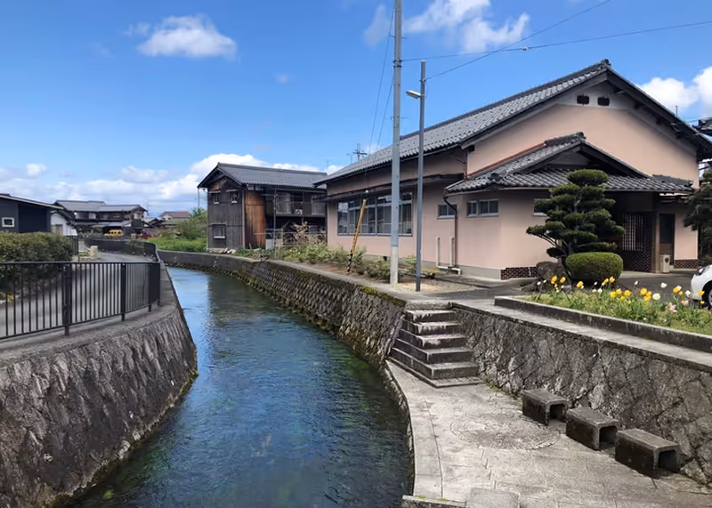 Clear canal running between stone walls with traditional Japanese-style houses and a blue sky with clouds overhead.