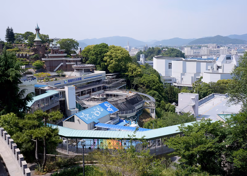 Himeji City Aquarium complex surrounded by trees with a castle-like structure nearby and a cityscape with mountains in the background.