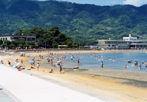 Crowded sandy beach with people swimming and relaxing, backed by lush green mountains and buildings.