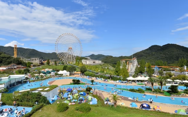 Wide view of an amusement park with a large Ferris wheel, water rides, green hills, and people enjoying pools and tents under a partly cloudy sky.