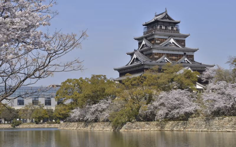 Traditional Japanese castle surrounded by cherry blossoms and greenery reflected in calm water under a clear blue sky.