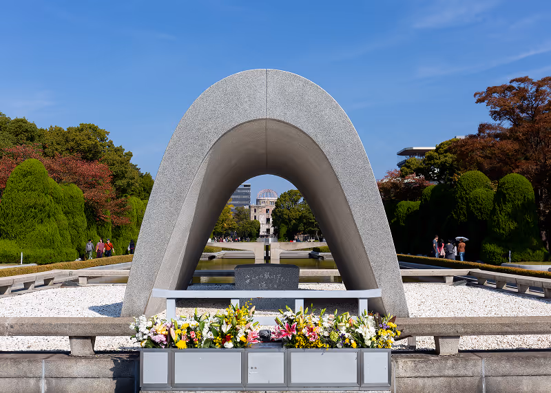 The Cenotaph for the A-bomb victims at Hiroshima Peace Memorial Park, a stone arch monument with colorful flowers in front and the Atomic Bomb Dome visible in the distance.