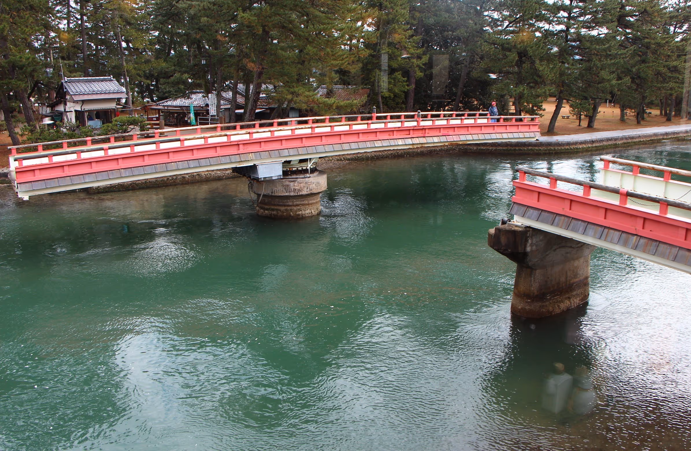 Red wooden bridge section lifted above green water with trees and small buildings in the background.
