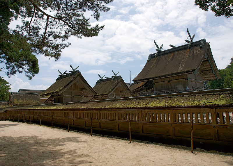 Traditional Japanese shrine buildings with large wooden roofs and moss-covered wooden fence under a partly cloudy sky.