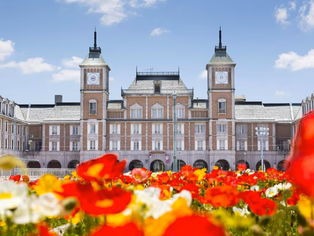 Brick building with two clock towers behind a colorful flower garden under blue sky.