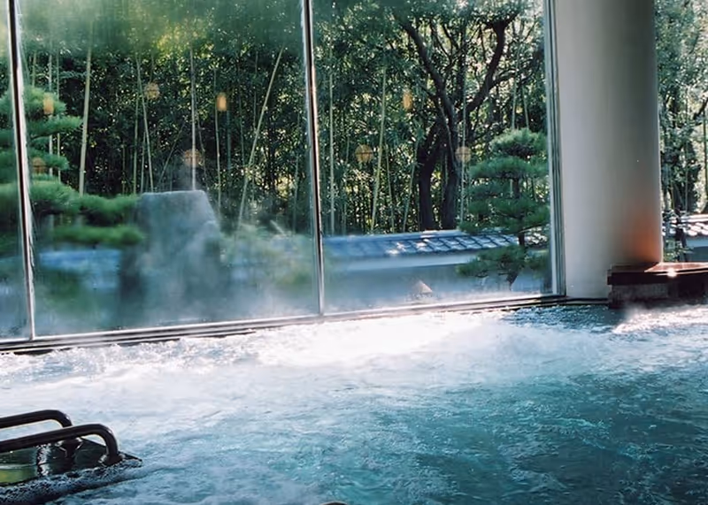 Indoor hot spring bath with bubbling water and large windows revealing a tranquil garden with green trees and bamboo.