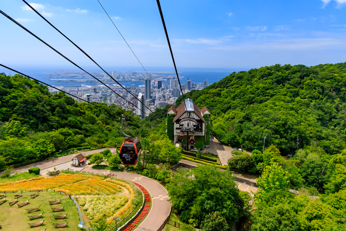 Cable cars on a ropeway descending through green hills with a cityscape and ocean in the background under a blue sky.