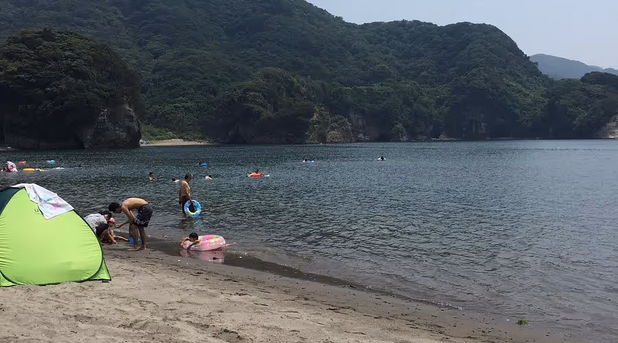 People enjoying a calm beach with green forested hills in the background, including children playing near the shore and a bright green tent on the sand.