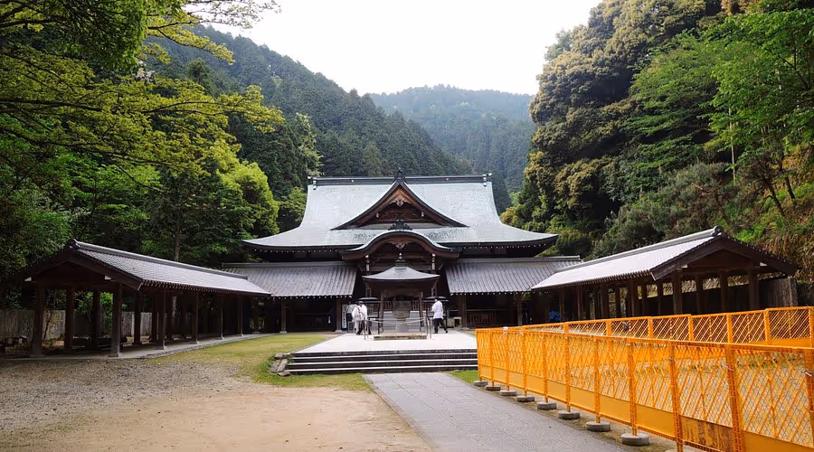 Traditional Japanese temple surrounded by lush green forest and mountains, with two people walking near the entrance.