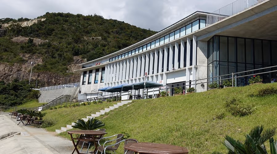 Modern building with large windows next to a grassy hill and outdoor seating, with a forested hill in the background under a cloudy sky.