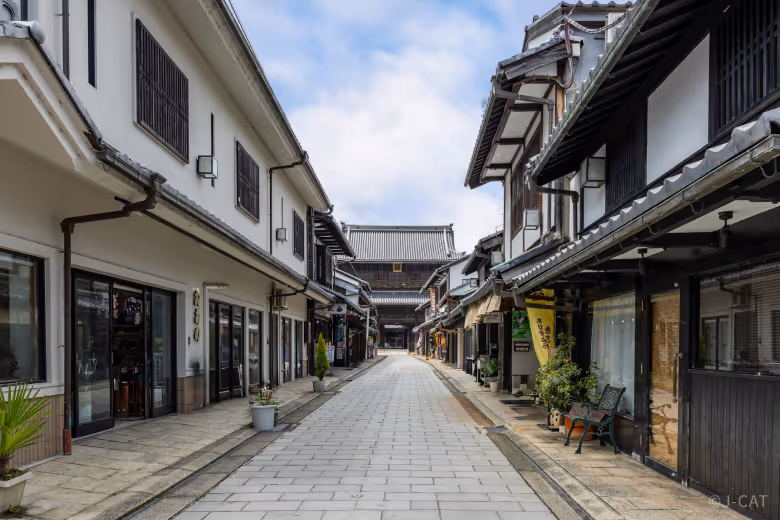 Narrow, paved street lined with traditional Japanese buildings and shops, leading to a historic gate under a partly cloudy sky.