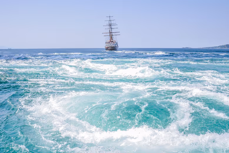 Sailing ship on calm blue sea with swirling whirlpools in the foreground.