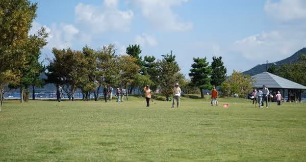 People walking and standing on a large grassy field with trees and a pavilion in the background under a partly cloudy sky.