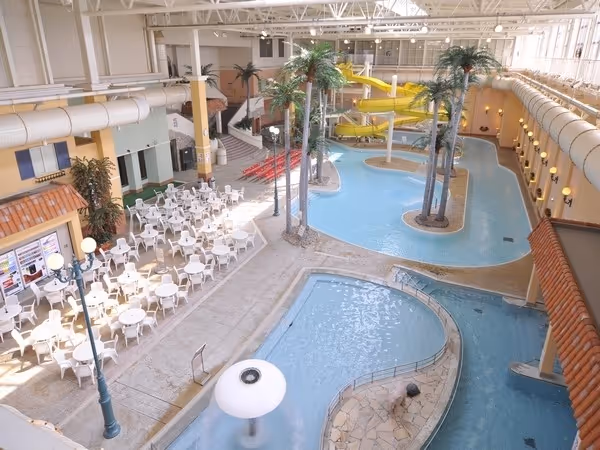 Indoor water park with winding pools, tall artificial palm trees, yellow water slides, and surrounding white chairs and tables.