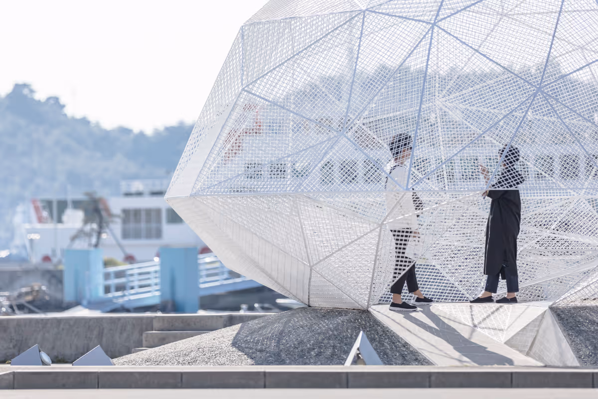 Two people standing and talking inside a large white geometric metal pavilion near a waterfront with boats and hills in the background.