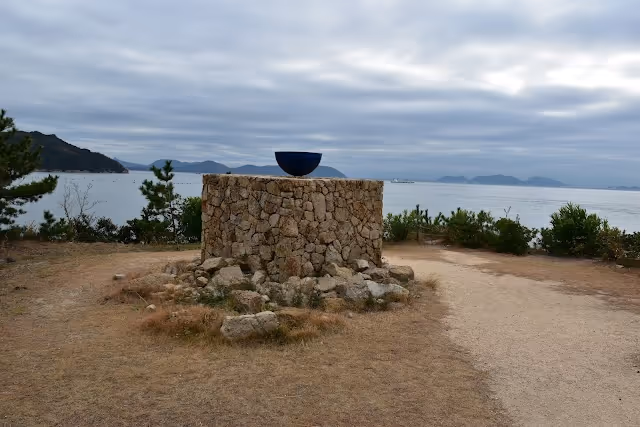 Stone pedestal with a dark bowl sculpture on top, set in a coastal landscape with cloudy sky and distant mountains over the water.