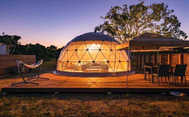 Illuminated geodesic dome tent on wooden platform at twilight with hammock and dining area outdoors.
