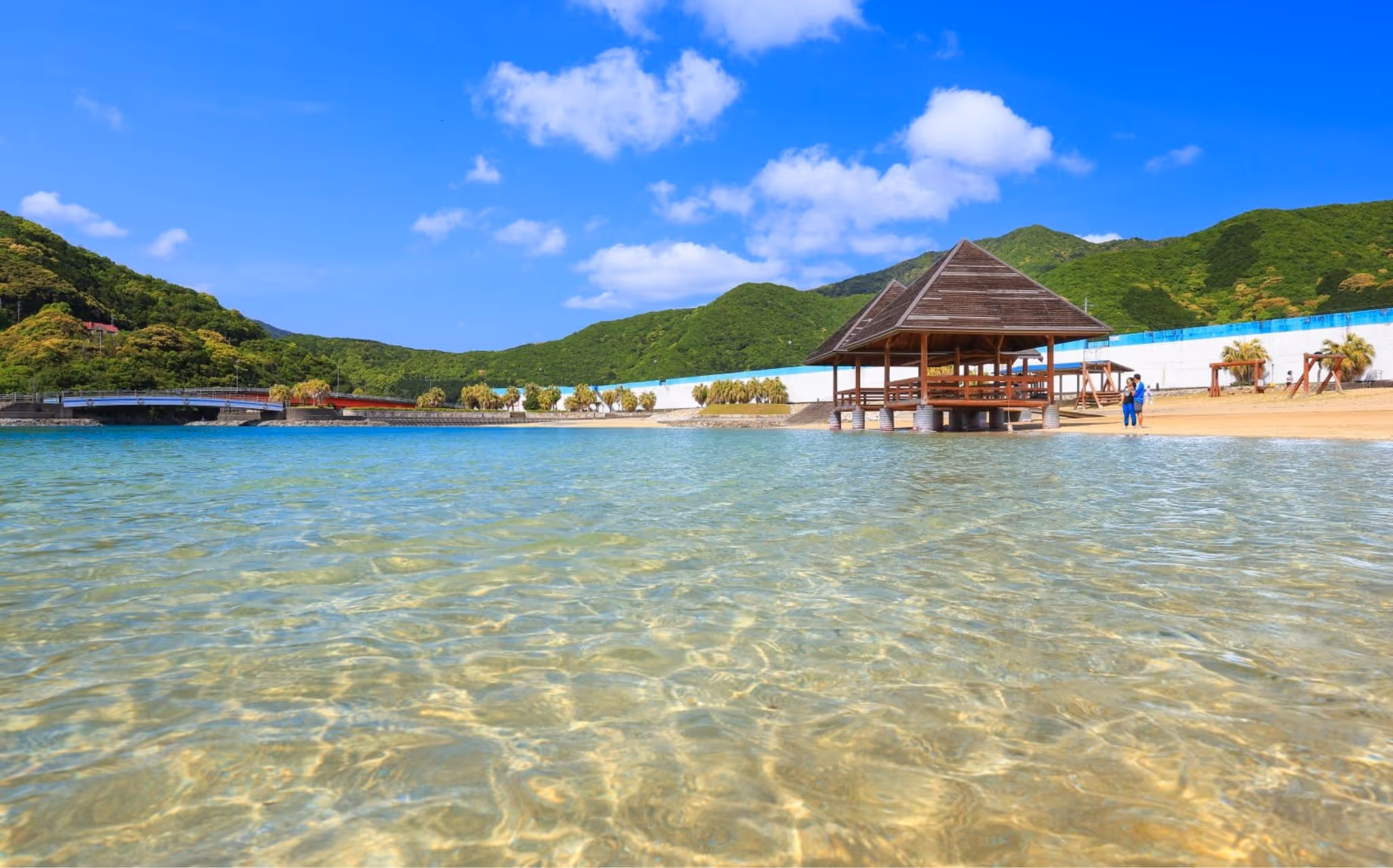 Clear shallow water with a wooden pavilion on stilts at the sandy shore, green hills, and blue sky with clouds in the background.