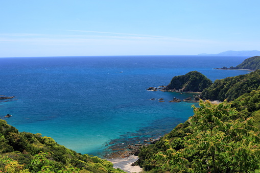 Clear blue ocean with rocky coastline and lush green hills under a bright sky.