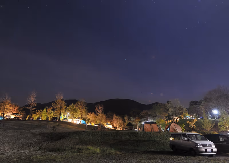 Nighttime view of a campground with illuminated tents and parked cars under a starry sky with distant mountains.