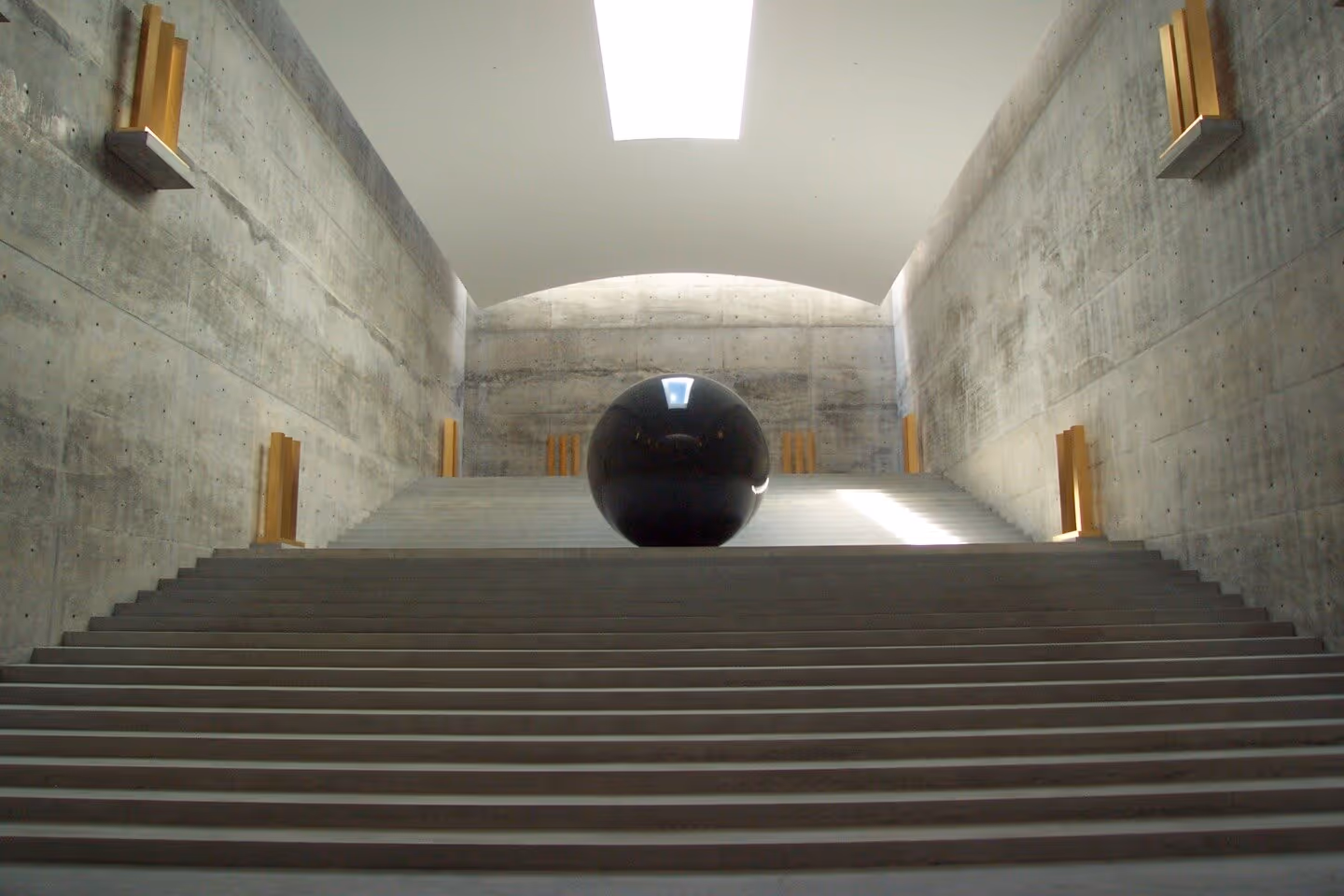 Large black reflective sphere centered on wide concrete stairs inside a minimalist room with concrete walls and a rectangular skylight.