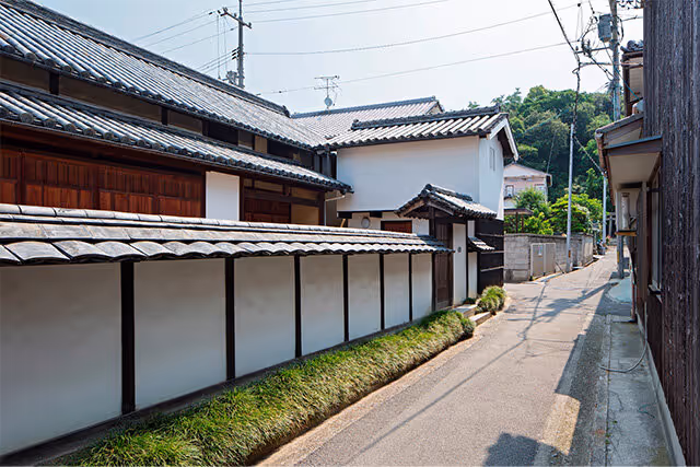 Narrow street lined with traditional Japanese buildings featuring white walls, wooden panels, and tiled roofs.