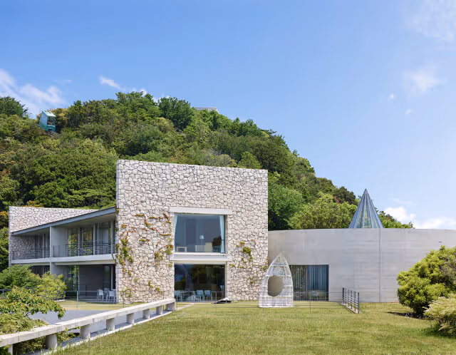 Modern stone and concrete building with large windows set in front of a green wooded hill under a blue sky.