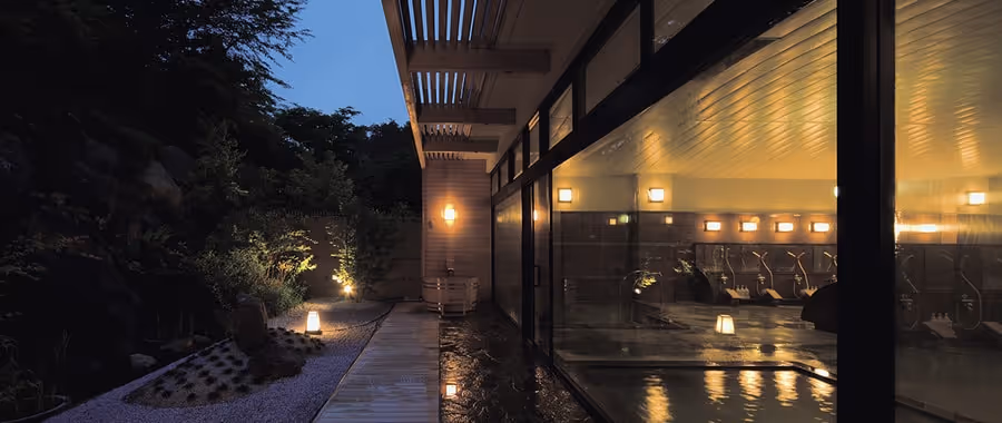 Evening view of a serene outdoor walkway next to a building with large glass windows revealing an illuminated indoor hot spring bath area.