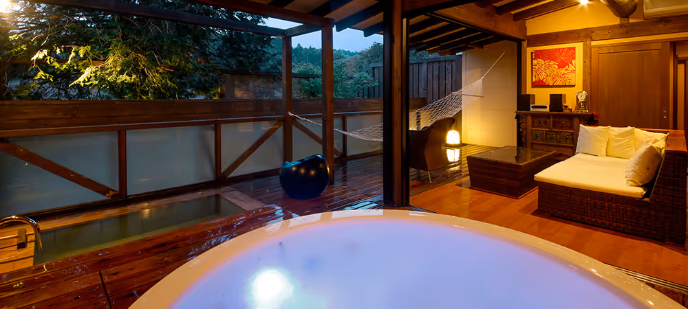 Cozy indoor-outdoor space with a lit hot tub in the foreground, wooden deck, hammock, and cushioned wicker seating under a roof.
