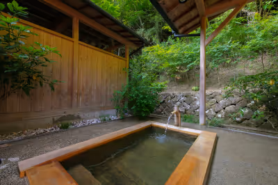 Outdoor wooden hot spring bath with natural stone retaining wall and lush greenery surrounding it.