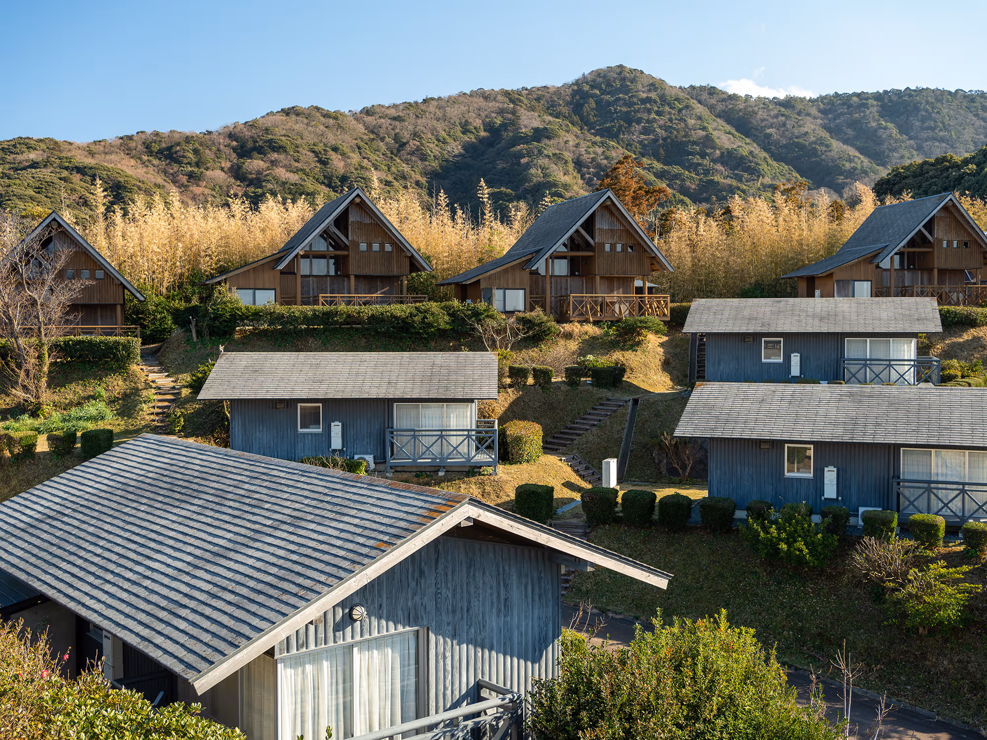 Wooden cabins and houses on a hillside with trimmed bushes, set against a backdrop of forested mountains under a clear blue sky.