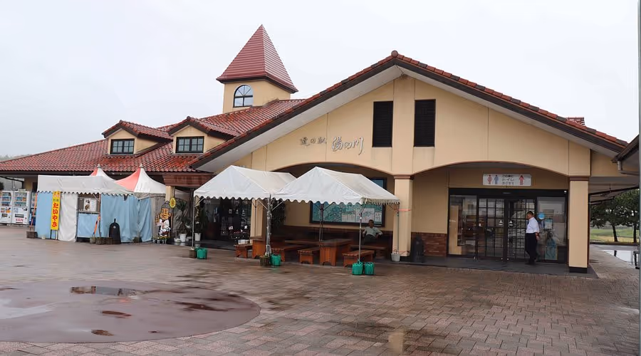 Building with a red tiled roof and a small tower, featuring tents and benches in front, with two people near the entrance on a cloudy day.