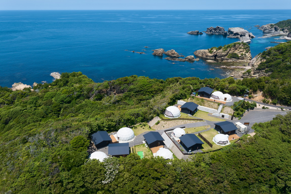 A coastal glamping site with white geodesic dome tents and black cabins surrounded by green trees overlooking a blue ocean with rocky formations.