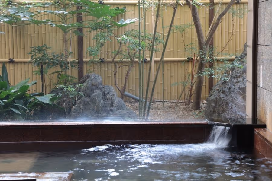 Indoor hot spring bath with water flowing from a spout, overlooking a garden with rocks, bamboo fence, and leafy plants.