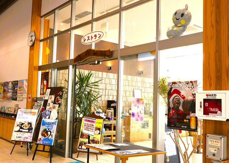 Glass entrance of a Japanese restaurant with menu boards, a large plant inside, and an AED emergency device mounted on the wooden wall.