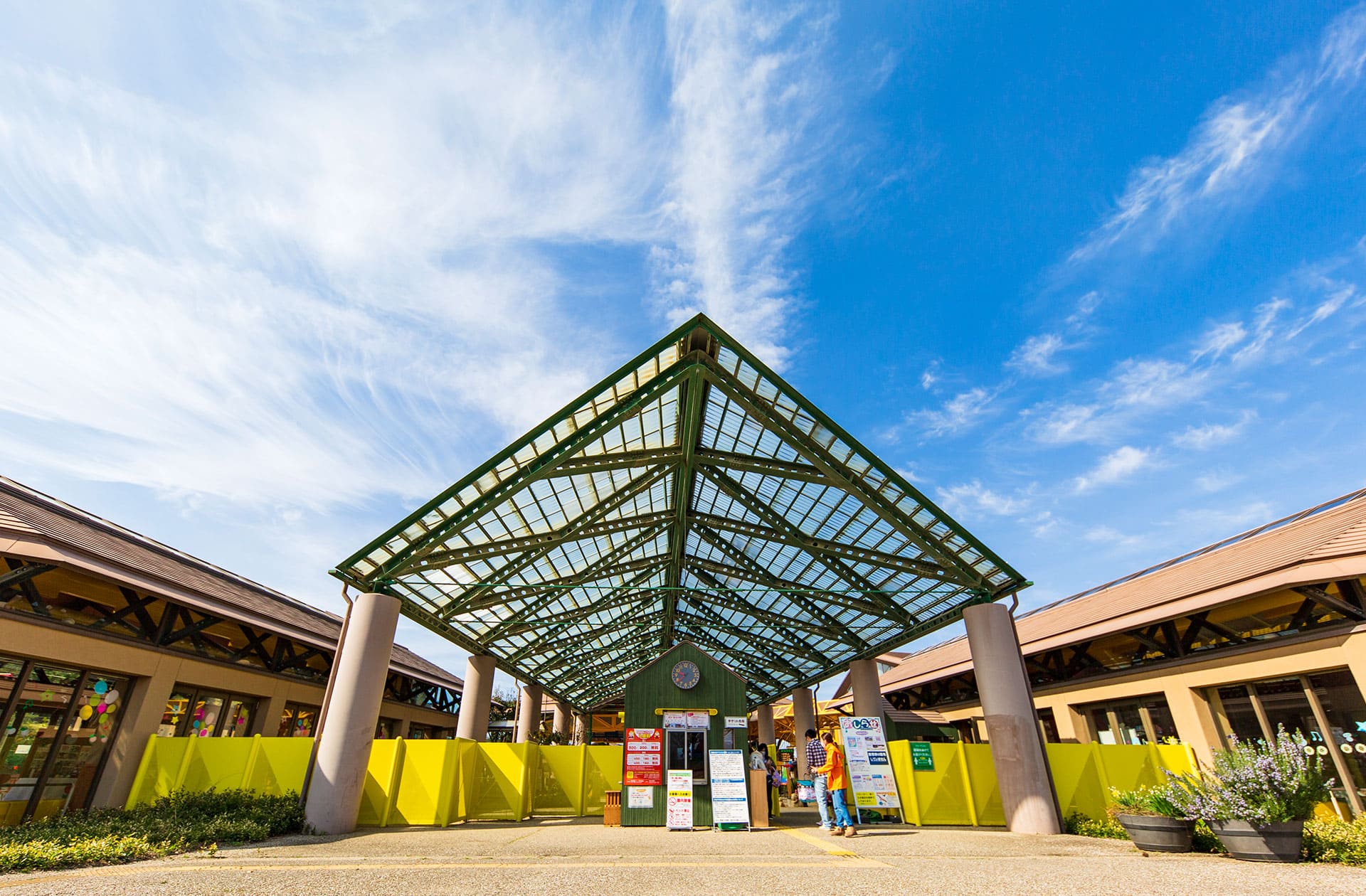 Outdoor entrance structure with green glass roof and yellow barriers under a bright blue sky.