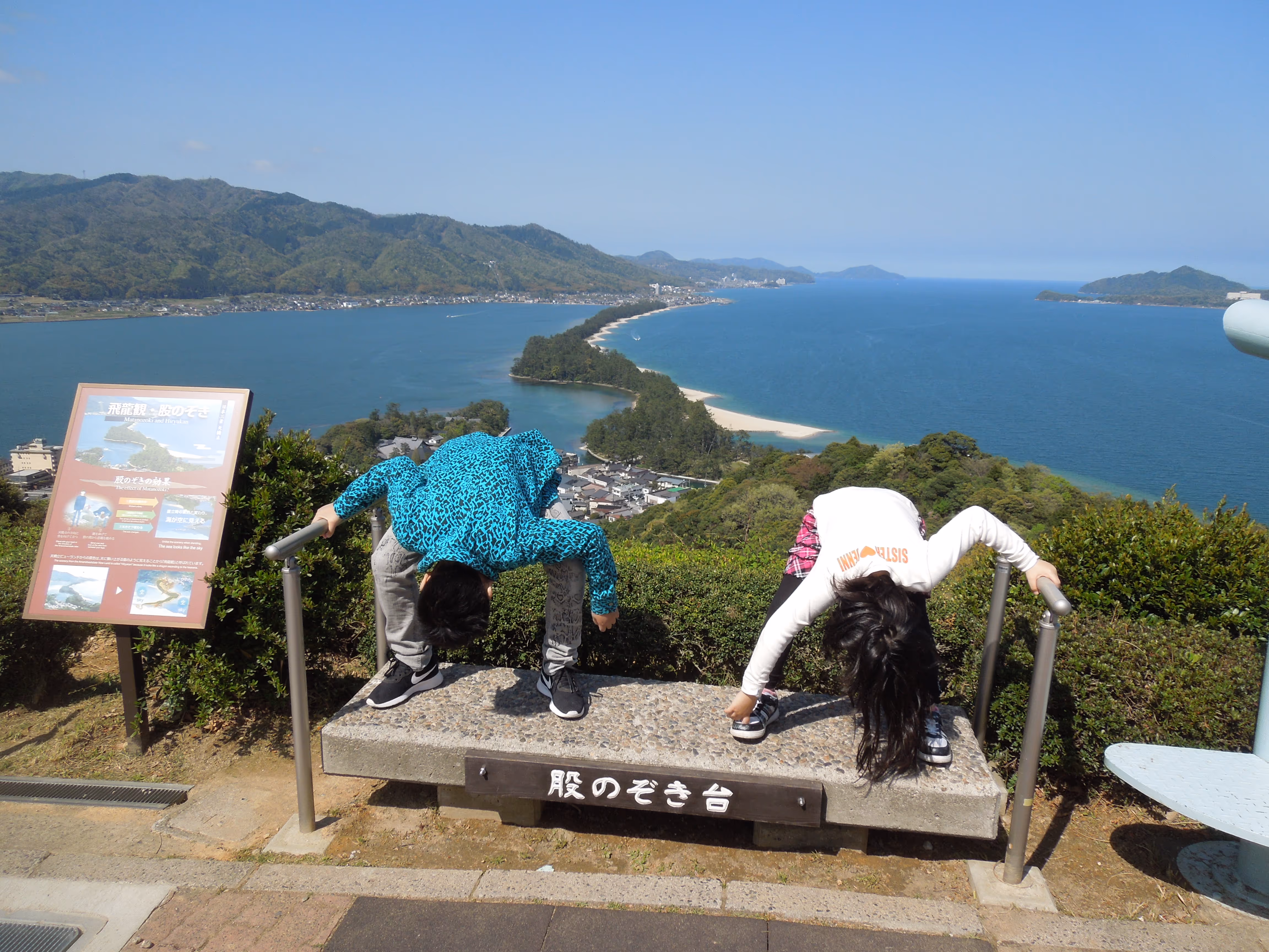 Two people leaning forward over a viewing platform railing overlooking a long sandbar extending into a blue sea with mountains in the background.